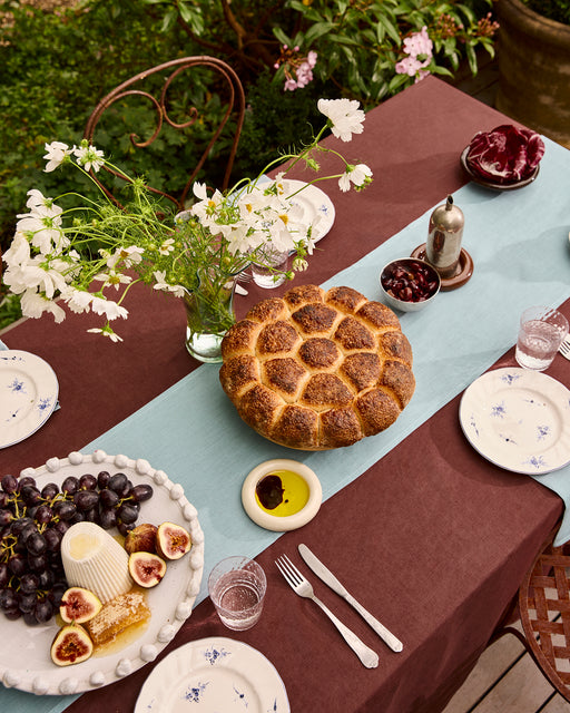 An outdoor meal is set with a brown tablecloth and a Lagoon 100% French Flax Linen Table Runner by Bed Threads, featuring bread, figs, grapes, cheese, placemats, white flowers in a vase, plates, silverware, and glasses.