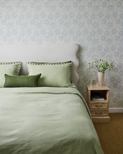 A neatly made bed with light green bedding and two pillows, including the Bed Threads Olive & Sage 100% French Flax Linen Piped Lumbar Cushion Cover, sits by a vintage nightstand with a glass of water and white flowers against pale blue patterned wallpaper. - view