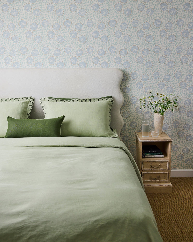 A neatly made bed with light green bedding and two pillows, including the Bed Threads Olive & Sage 100% French Flax Linen Piped Lumbar Cushion Cover, sits by a vintage nightstand with a glass of water and white flowers against pale blue patterned wallpaper.