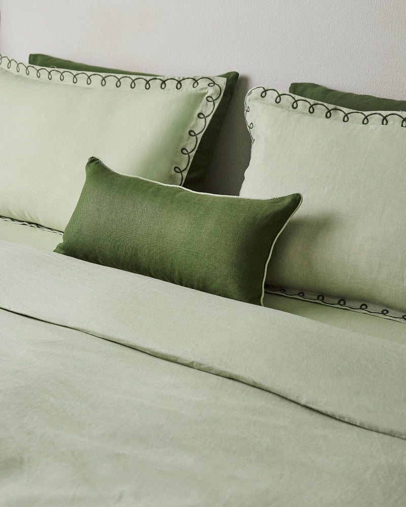 A neatly made bed with sage green bedding, two light green pillows with dark green piping, and a Bed Threads Olive & Sage 100% French Flax Linen Piped Lumbar Cushion Cover in dark green in front.