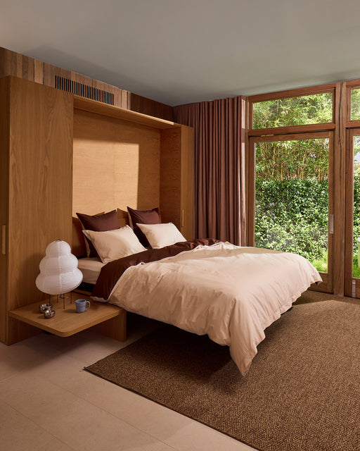 A modern bedroom features a wooden bed frame, Bed Threads’ Espresso, Vanilla & Sky Piped 100% Organic Cotton Duvet Cover, brown and white pillows, curtained glass doors, a brown rug, a garden view, plus a lamp and mugs on the bedside shelf.