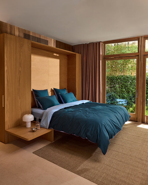 A modern bedroom with a wooden Murphy bed, teal bedding, Bed Threads Aegean, Sky & Bubblegum Piped 100% Organic Cotton European Pillowcases (Set of Two), two bedside tables with lamps, floor-to-ceiling windows, beige curtains, and greenery views.