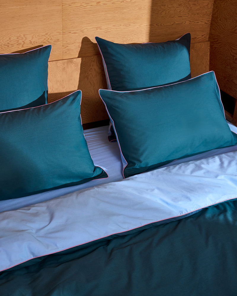 A neatly made bed with teal pillows, a matching duvet, and light blue sheets is accented by Bed Threads' Aegean, Sky & Bubblegum Piped 100% Organic Cotton European Pillowcases (Set of Two), set against a wooden wall for a modern, cozy feel.