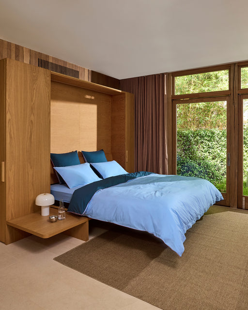 A modern bedroom with a wooden Murphy bed pulled down, dressed in blue bedding and Bed Threads’ Aegean, Sky & Bubblegum Piped 100% Organic Cotton European Pillowcases. A nightstand with a lamp and glassware sits by large windows framing lush greenery.