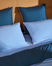 A bed with matching bedding features two Aegean and two Sky & Bubblegum Piped 100% Organic Cotton European Pillowcases by Bed Threads, all with contrasting trim, arranged neatly against a wooden headboard. - view