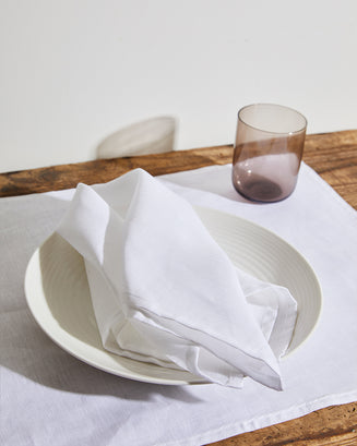 The Sage & White Table Bundle by Bed Threads features a white napkin on a matching plate atop a wooden table with a white placemat, accompanied by an empty translucent glass tumbler for an elegant dining setup.