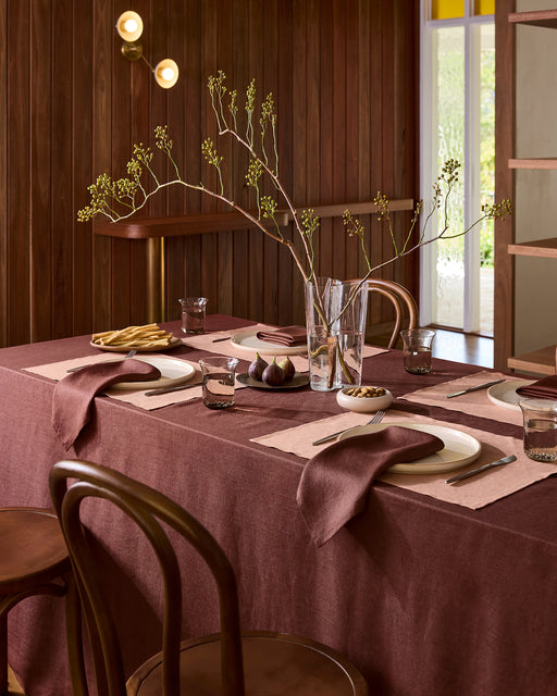 A table set for two features Bed Threads Hazelnut 100% French Flax Linen Placemats, white plates, burgundy napkins, silver cutlery, and water glasses. A bowl of nuts and a vase with branches add charm atop a dark burgundy tablecloth.