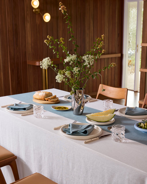A dining room with wood-paneled walls and floor features a Bed Threads White & Lagoon 100% French Flax Linen Table Bundle on the table, set for a meal with dishes and glassware. Two wooden chairs are placed at the table.