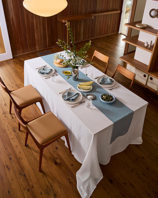 A Lagoon 100% French Flax Linen Table Runner by Bed Threads adorns a rustic outdoor table on a wooden deck, set for dining amid lush greenery and flowers, with tableware and string lights creating an inviting atmosphere.