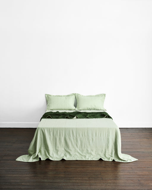 A neatly made bed with the Bed Threads Sage & Olive Two-Tone Quilt sits against a plain white wall on a dark wooden floor, featuring light green tones and matching pillows for a cohesive look.
