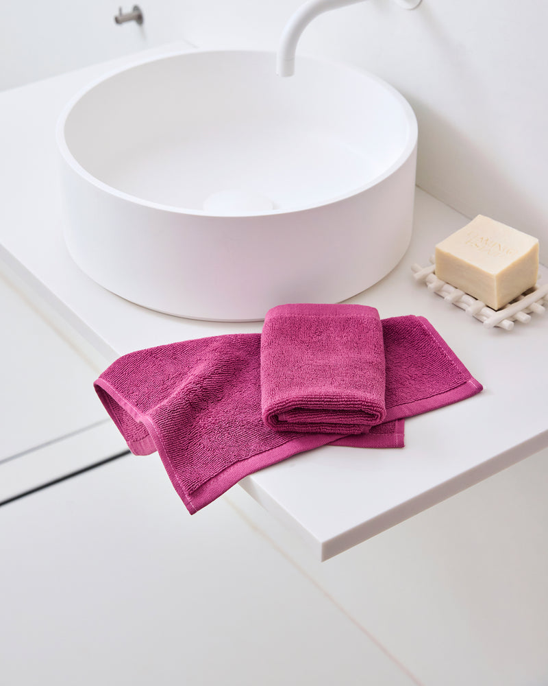 A modern white bathroom sink features Bed Threads Berry 100% Cotton Terry Face Washers (Set of Two) and a magenta towel neatly on the counter. A bar of soap on a dish beside the sink completes the clean, minimalistic look.
