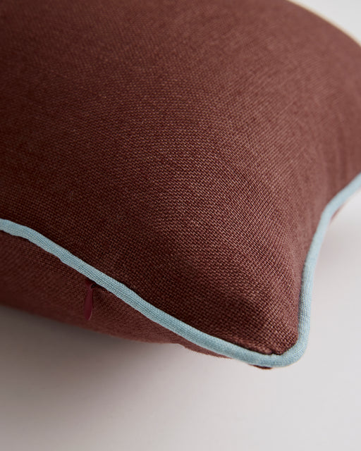 Close-up of the Bed Threads Cacao & Lagoon 100% French Flax Linen Lumbar Cushion Cover, featuring brown fabric with light blue contrast piping, resting on a white surface.