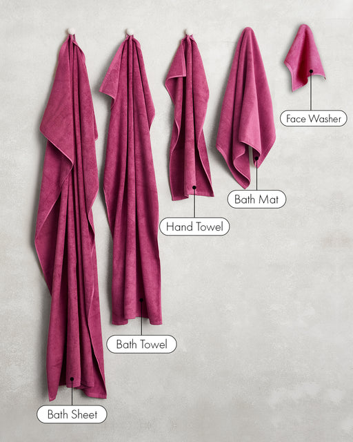 Five magenta towels, including the Berry 100% Cotton Terry Bath Mat by Bed Threads, hang on hooks against a light wall, labeled left to right: Bath Sheet, Bath Towel, Hand Towel, Bath Mat, and Face Washer.