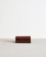 The Bed Threads Espresso 100% Cotton Terry Face Washers (Set of Two) feature dark brown rectangles with stitched edges and fabric labels, layered on a light surface. Made from absorbent terry, they're gentle on skin. - view