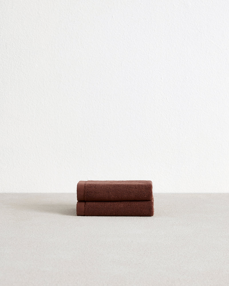 The Bed Threads Espresso 100% Cotton Terry Face Washers (Set of Two) feature dark brown rectangles with stitched edges and fabric labels, layered on a light surface. Made from absorbent terry, they're gentle on skin.