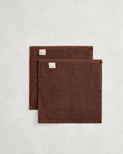 The Bed Threads Espresso 100% Cotton Terry Face Washers (Set of Two) feature dark brown rectangles with stitched edges and fabric labels, layered on a light surface. Made from absorbent terry, they're gentle on skin. - view