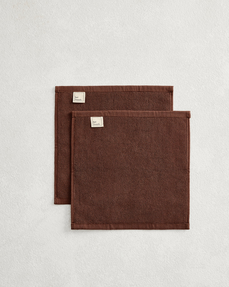 The Bed Threads Espresso 100% Cotton Terry Face Washers (Set of Two) feature dark brown rectangles with stitched edges and fabric labels, layered on a light surface. Made from absorbent terry, they're gentle on skin.