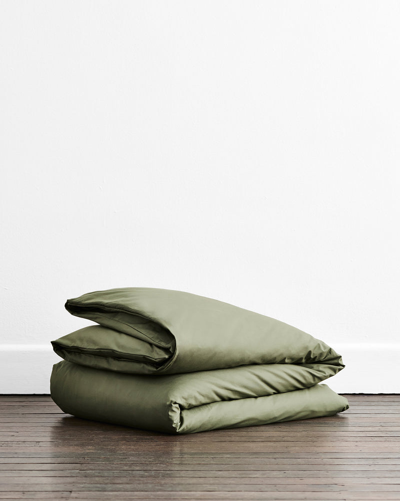 An olive green folded duvet cover in 100% organic cotton, on a dark wooden floor against a plain white wall.
