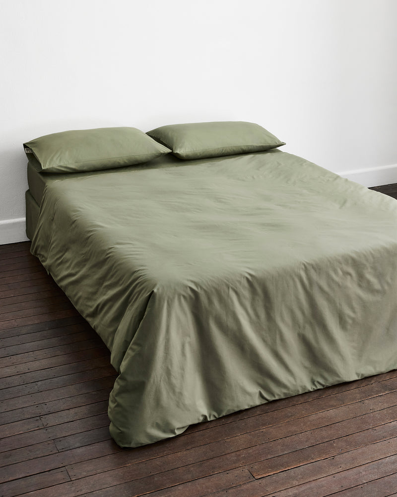 An olive green bed, including a duvet cover and two matching pillowcases in 100% organic cotton, styled on a bed against a white wall and dark wood floor.