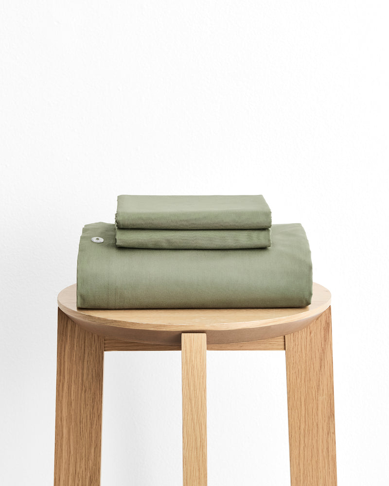 A folded stack of sage green bedding, including a duvet cover and two pillowcases displayed on a light wood stool against a plain white background.