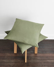 Two sage green european pillows in organic cotton arranged on a wooden stool against a white wall and dark wood floor. - view