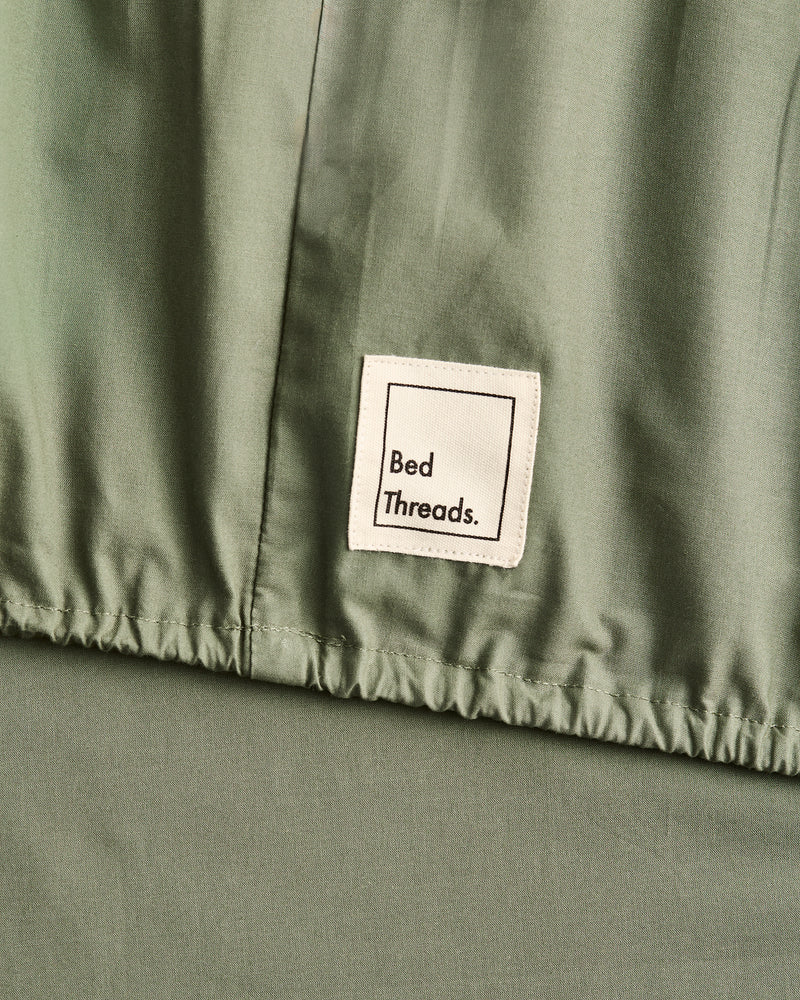 Close-up of a sage green fitted sheet elastic with a white sewn-on label reading 'Bed Threads.'