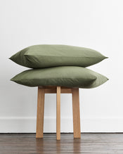 Two olive green organic cotton standard pillowcases stacked on a light wood stool against a white wall and dark wood floor. - view