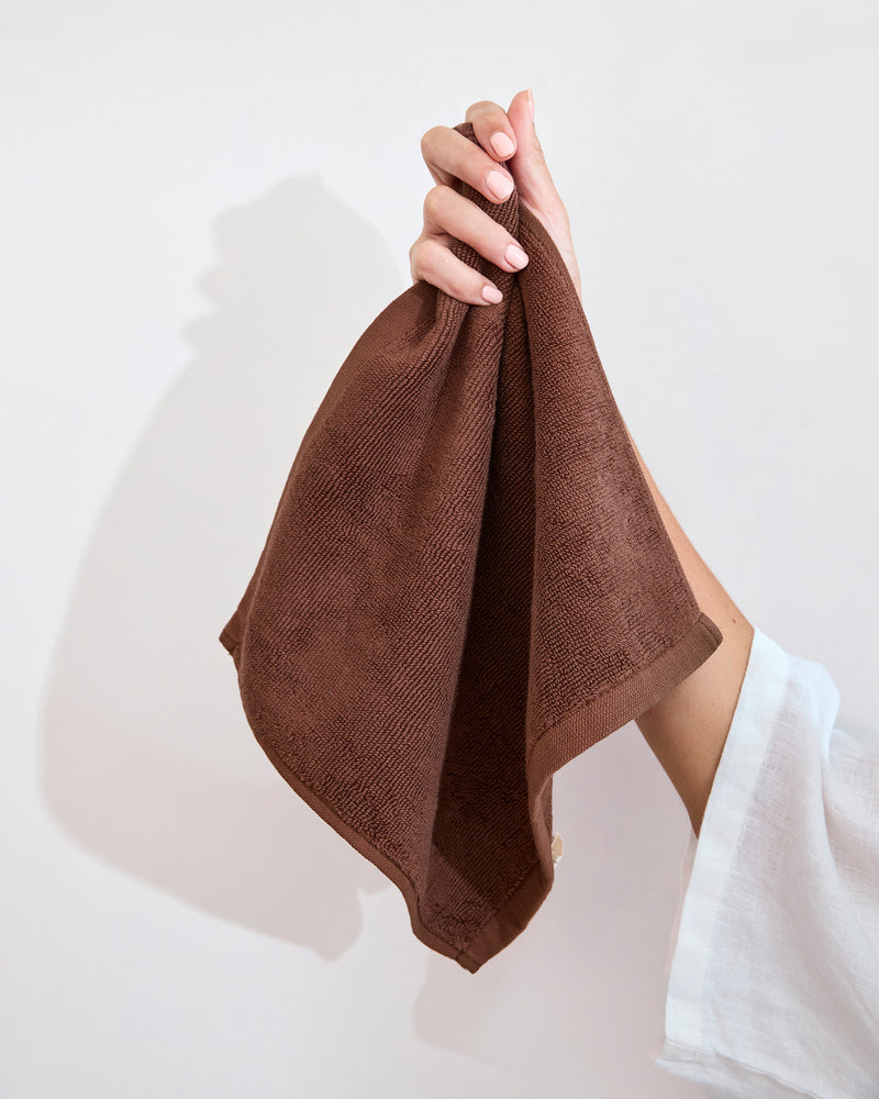 A folded Espresso 100% Cotton Terry Face Washer by Bed Threads sits on a white bathroom countertop beside a round sink and soap dish. The absorbent, gentle terry adds comfort and style to the modern, minimal setting. Set of two.