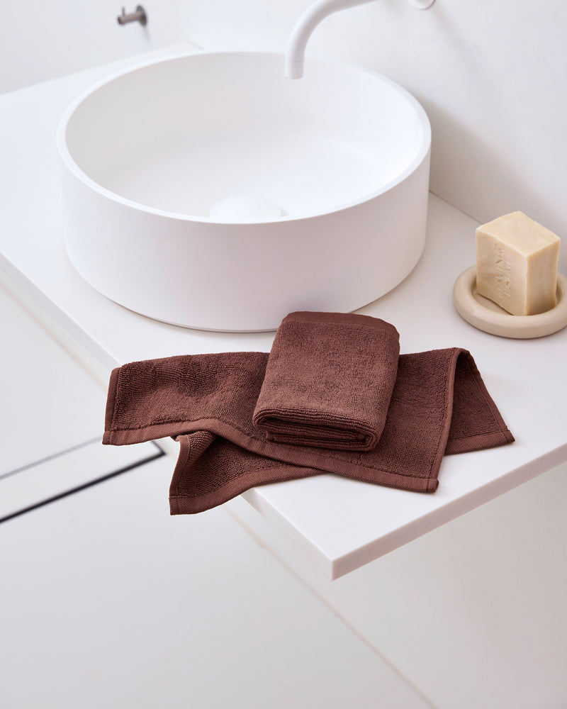A folded Espresso 100% Cotton Terry Face Washer by Bed Threads sits on a white bathroom countertop beside a round sink and soap dish. The absorbent, gentle terry adds comfort and style to the modern, minimal setting. Set of two.