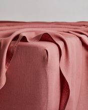 A close-up of a bed corner shows the Bed Threads Pink Clay 100% French Flax Linen Fitted Sheet, softly draped and folded over the edge against a plain, light background. - view