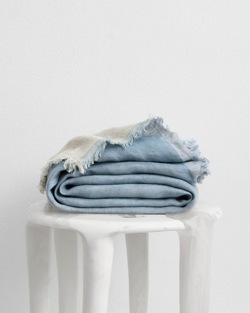 The Lagoon & Crème 100% French Flax Linen Muslin Throw by Bed Threads, in blue and beige, is neatly folded atop a smooth white marble stool against a plain white wall.
