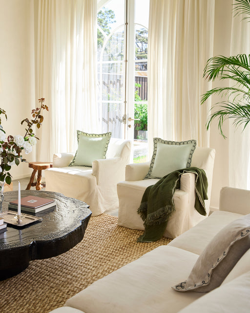 A light armchair with a Bed Threads Sage & Olive 100% French Flax Linen Swirl Cushion Cover and a dark green throw, set on a woven rug by a window with sheer curtains and greenery outside.