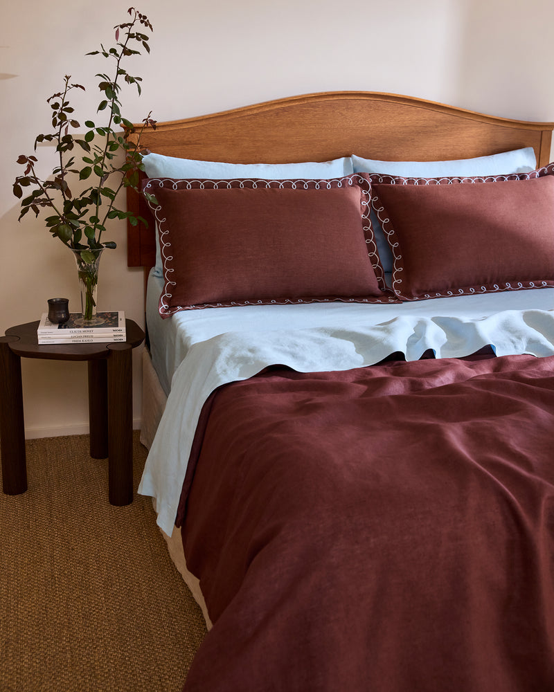 A neatly made bed with a wooden headboard features light blue sheets and brown bedding, topped with Bed Threads' Cacao & Coast 100% French Flax Linen Swirl Pillowcases. Nearby, a round table holds a vase with branches and a stack of books.