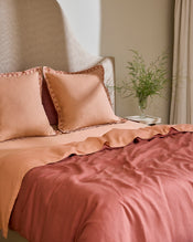 A bed styled with Bed Threads Terracotta & Pink Clay 100% French Flax Linen Swirl European Pillowcases, a beige upholstered headboard, and a side table topped with a glass vase of green foliage. - view