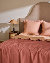 A neatly made bed features Bed Threads’ Terracotta & Pink Clay 100% French Flax Linen Swirl European Pillowcases (Set of Two), beside a small round wooden nightstand with a stone-based lamp and woven lampshade against a light wall. - view