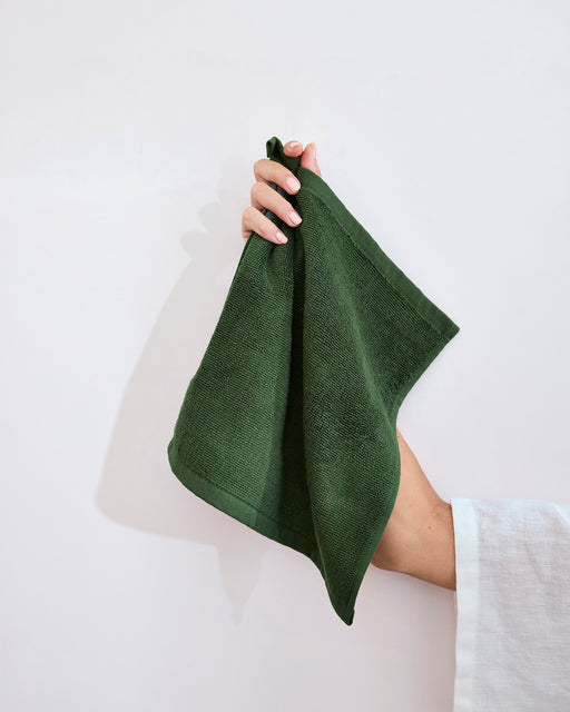 A modern white bathroom sink features a folded dark green towel and Bed Threads Moss 100% Cotton Terry Face Washers (Set of Two) on the counter, with a beige sponge in a wooden dish creating an inviting daily care space.