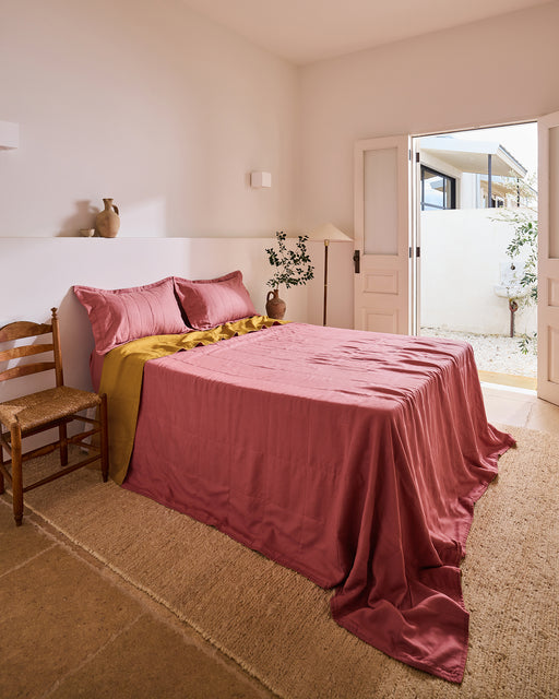 A cozy bedroom features a bed topped with Bed Threads' Pink Clay & Turmeric Two-Tone Quilt, pink pillows, a wooden chair, a vase on a shelf, beige walls, a woven rug, and open double doors revealing an outdoor area with white walls.