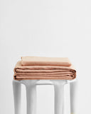 A neatly folded stack of beige or light brown bedsheets sits on top of a white stool against a plain white background.