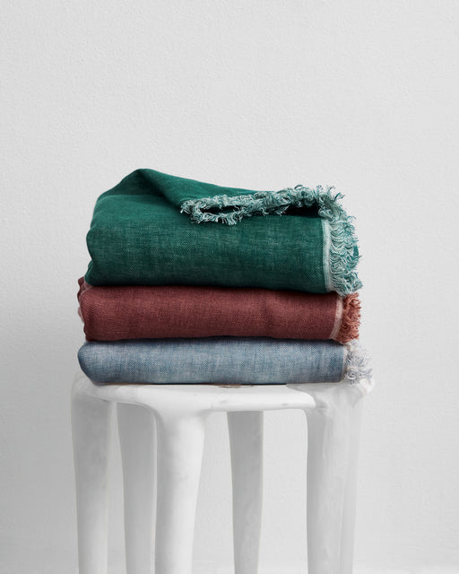 A stack of three Bed Threads Cacao & Hazelnut 100% French Flax Linen Muslin Throws, in green, reddish-brown, and blue, is folded on a white minimalist stool against a plain white wall.