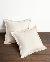 Two Crème & Coast 100% French Flax Linen Swirl European Pillowcases by Bed Threads, set of two, in beige with light blue decorative edge stitching and zip closure, displayed on a wooden floor against a white wall. - view