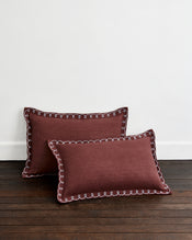 Two Cacao & Coast 100% French Flax Linen Swirl Pillowcases by Bed Threads in maroon, with white decorative stitching, are placed on a dark wooden floor against a plain white wall. - view