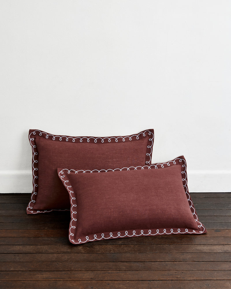 Two Cacao & Coast 100% French Flax Linen Swirl Pillowcases by Bed Threads in maroon, with white decorative stitching, are placed on a dark wooden floor against a plain white wall.