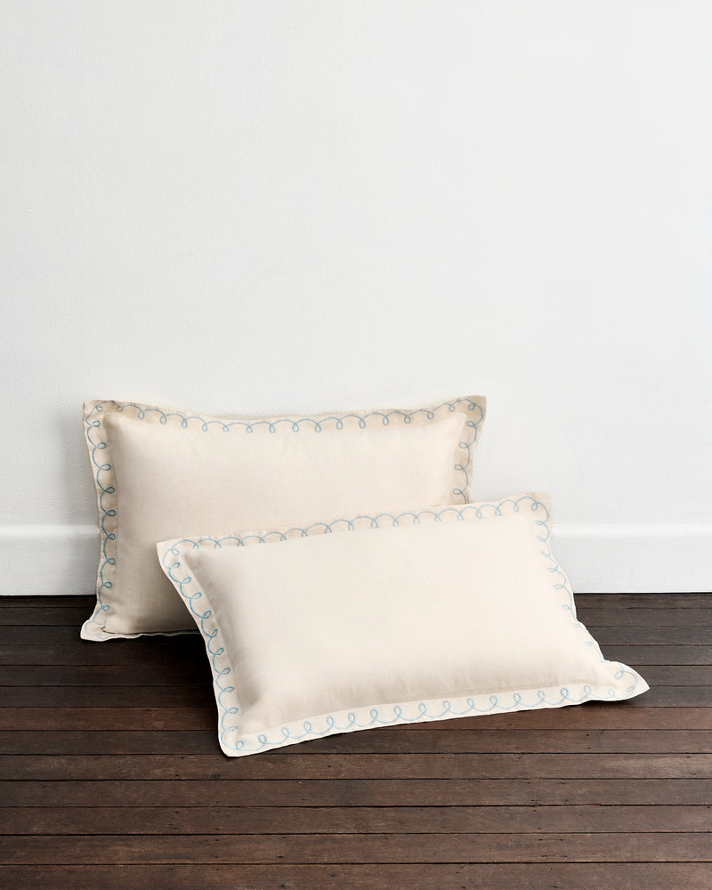 Two Crème & Coast 100% French Flax Linen Swirl Pillowcases by Bed Threads rest against a white wall on a dark wooden floor, featuring elegant embroidered edges and offering the perfect upgrade for breathable linen bedding.
