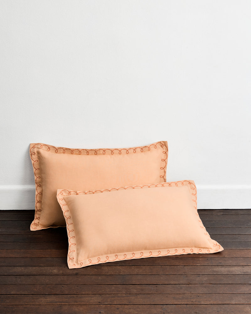 The Bed Threads Terracotta & Pink Clay 100% French Flax Linen Swirl Pillowcases (Set of Two) showcase elegant, breathable linen and swirl-embroidered edges, displayed against a dark wooden floor and a plain white wall.