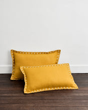 Two Turmeric & Crème 100% French Flax Linen Swirl Pillowcases by Bed Threads are placed on a dark wooden floor, leaning against a white wall. - view