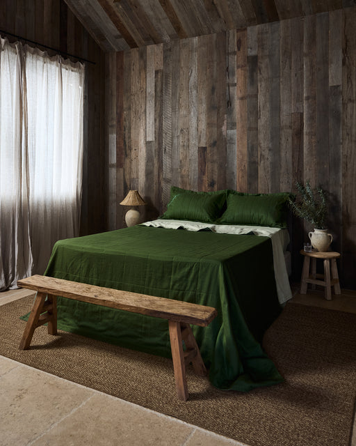 A rustic bedroom with wooden walls, a bed topped with Bed Threads' Sage & Olive Linen Quilt, two pillows, a wooden bench at the foot, a woven rug, and a side table with a ceramic jug and greenery. Sunlight filters through sheer curtains.