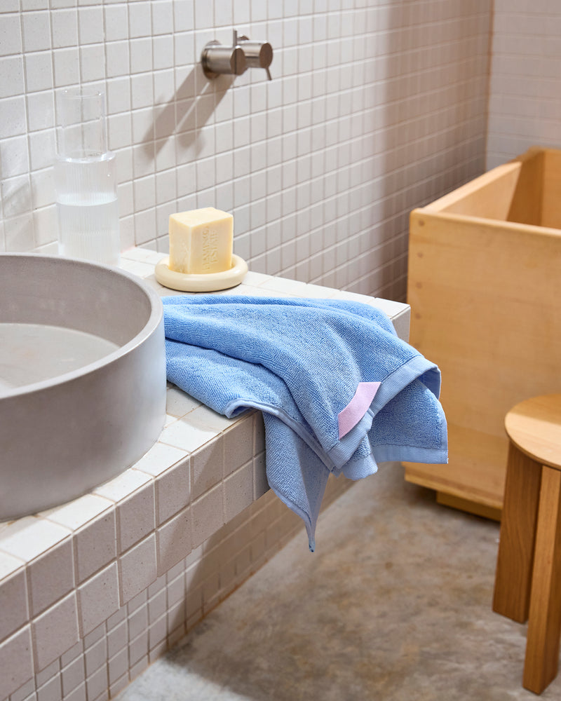 A Bed Threads Sky 100% Cotton Terry Hand Towel, a bar of soap on a dish, and a glass of water sit on a white tiled bathroom sink beside a round basin, with a wooden stool and tub in the background.
