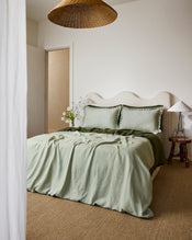A bed dressed in light green and olive tones features Bed Threads’ Sage & Olive 100% French Flax Linen Swirl Pillowcases, two large swirl-edged pillows, a curved upholstered headboard, and a rustic wooden side table with a white lamp. - view