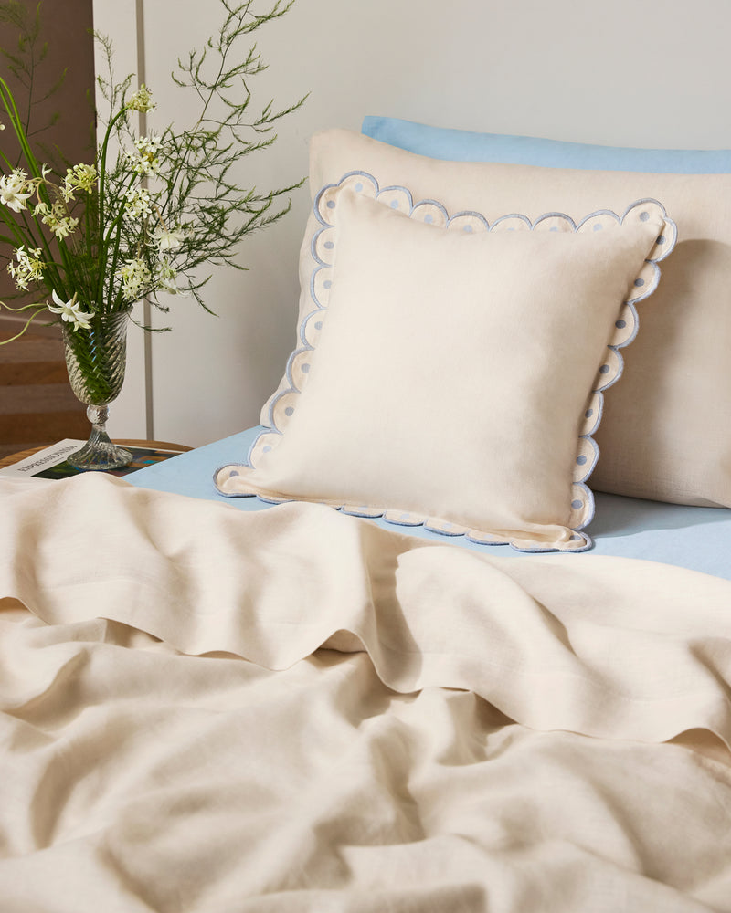 A neatly made bed with Bed Threads' Crème & Coast 100% French Flax Linen Arch Cushion Cover, light blue sheets, a matching cream blanket, and a glass vase with white flowers and greenery on the bedside table.
