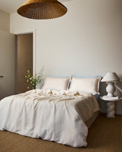 Two Crème & Coast 100% French Flax Linen Swirl Pillowcases by Bed Threads, in cream with blue embroidered edges, rest against a light blue headboard and bedding. The soft, breathable linen appears slightly wrinkled for a relaxed look. - view